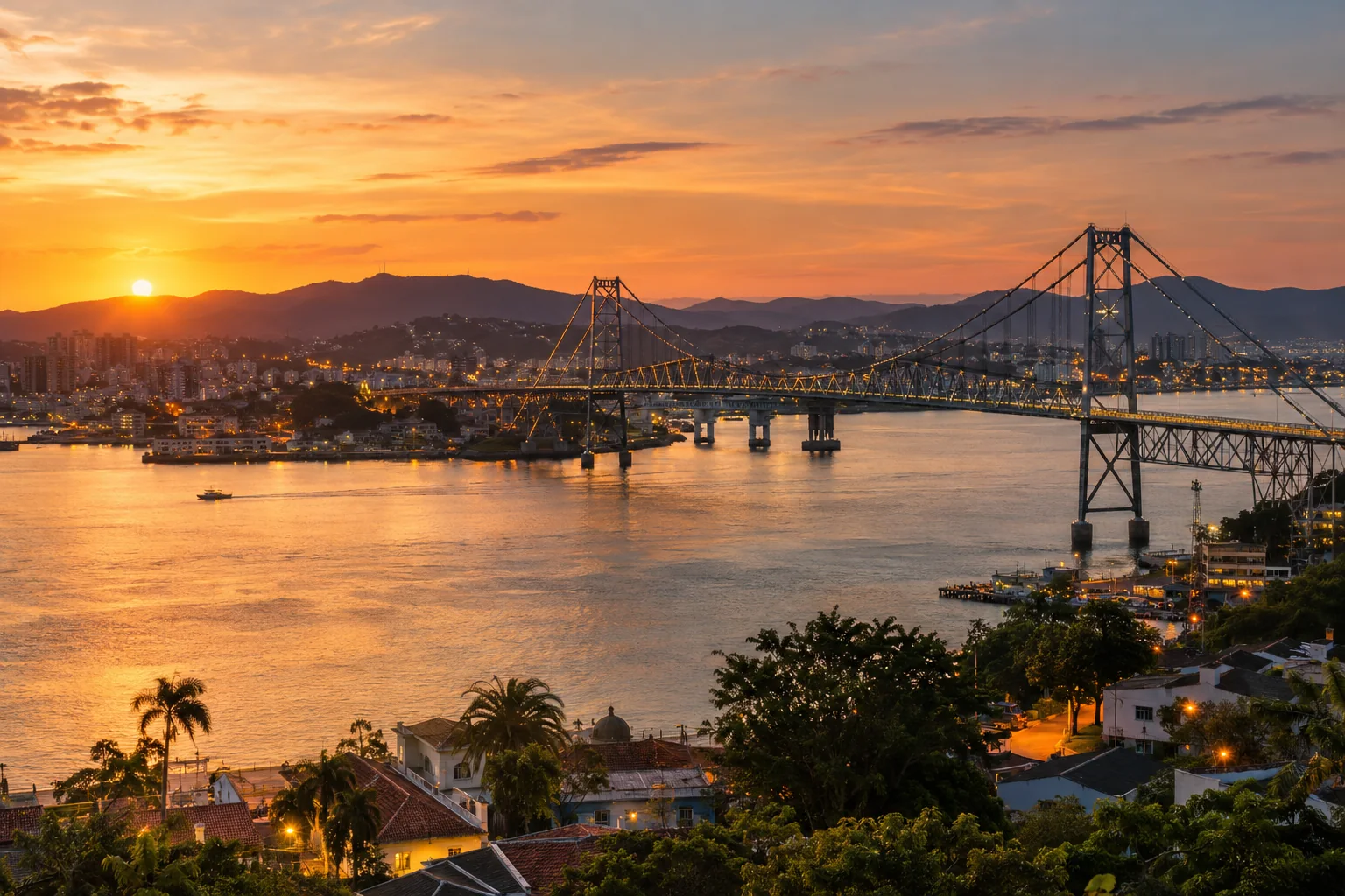 View over the waterfront and the distinctive Hercílio Luz suspension bridge, coastal hills and city lights beginning at warm dusk